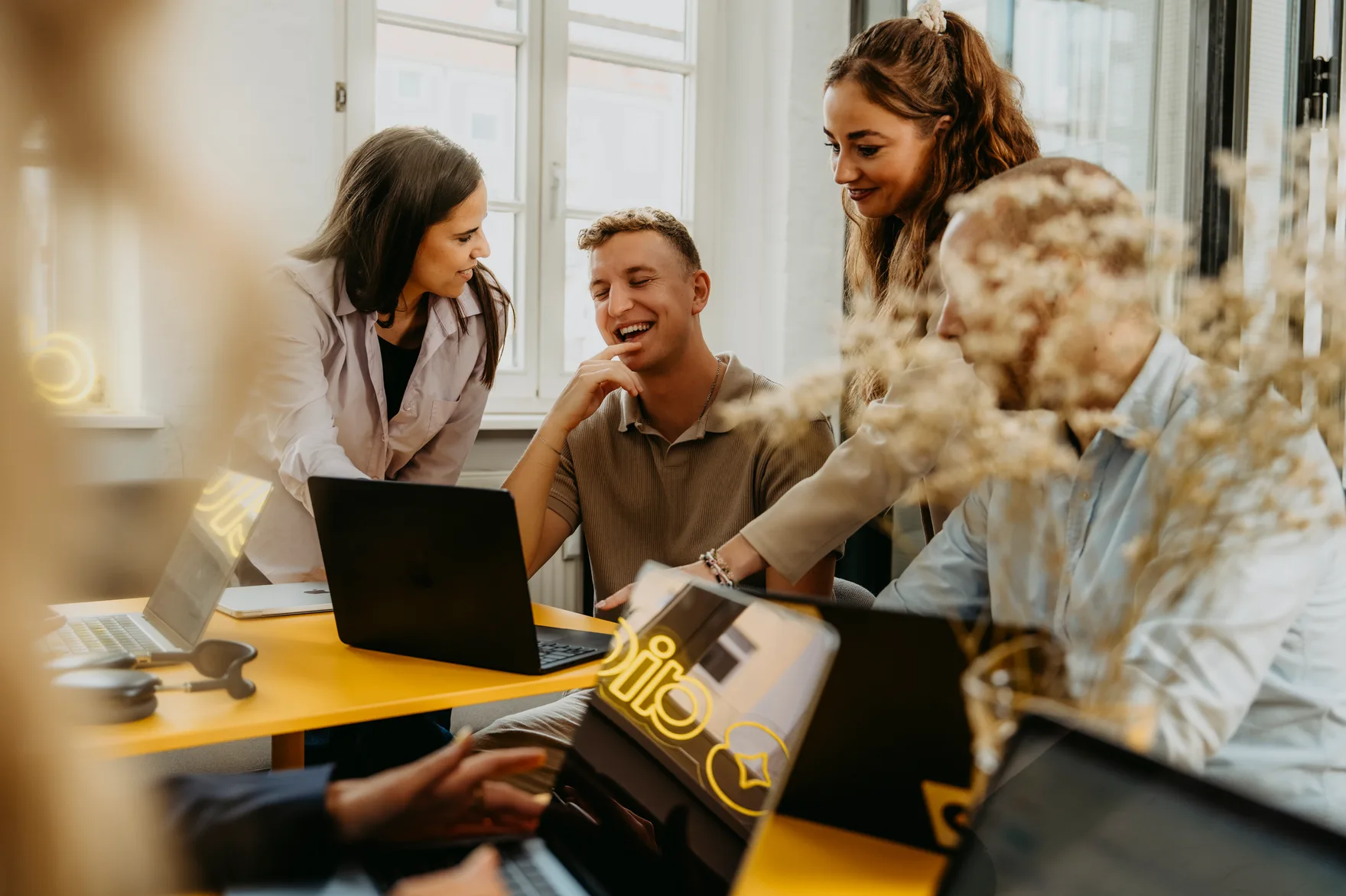 Vier Personen lachen gemeinsam an einem gelben Tisch mit Laptops